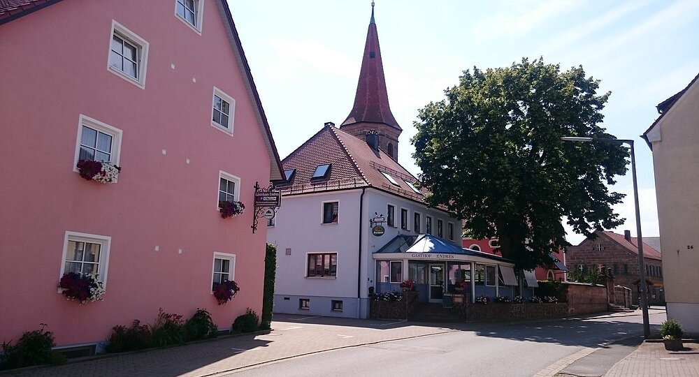 Link am Rand ein großes rosanene Haus. Daneben etwas weiter im Hintergrund ein grauer Gasthof mit Sitzgelegenheiten und Pavillon direkt vor dem Haus. Links folgt ein großer Baum.