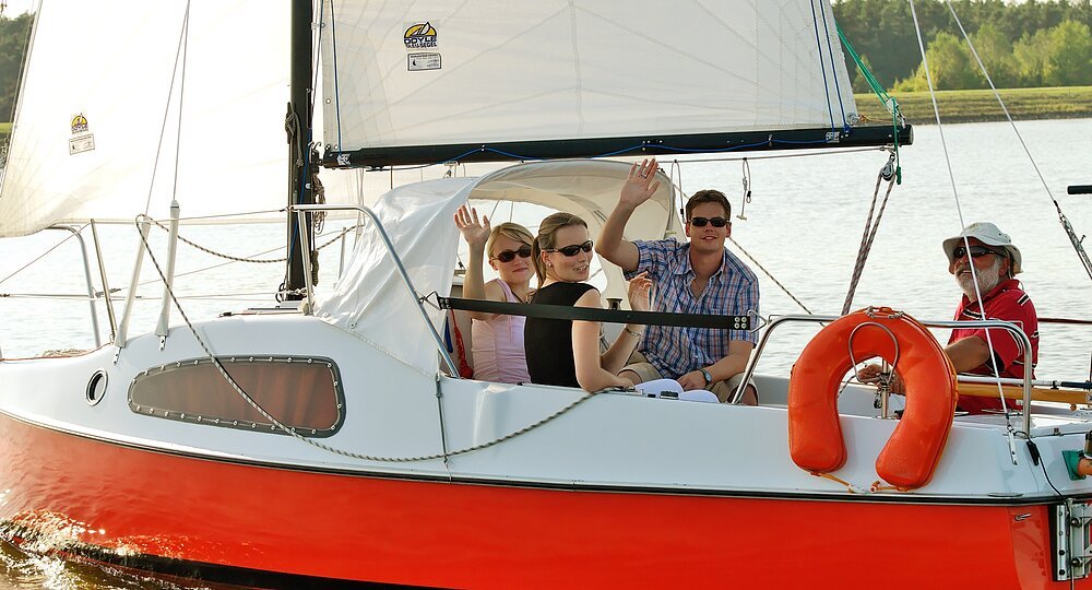 Zwei Frauen und ein Mann sitzen auf einem rot weißen Segelboot und winken fröhlich in die Kamera. Ein älterer Mann steuert das Boot über den Rothsee.