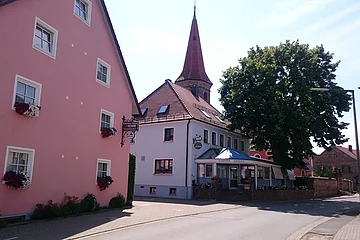 Link am Rand ein großes rosanene Haus. Daneben etwas weiter im Hintergrund ein grauer Gasthof mit Sitzgelegenheiten und Pavillon direkt vor dem Haus. Links folgt ein großer Baum.