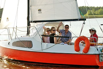 Zwei Frauen und ein Mann sitzen auf einem rot weißen Segelboot und winken fröhlich in die Kamera. Ein älterer Mann steuert das Boot über den Rothsee.